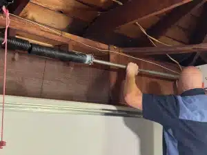 A service technician works on a metal torsion shaft assembly mounted against a wooden header beam, demonstrating the complex process of how to replace garage door springs.