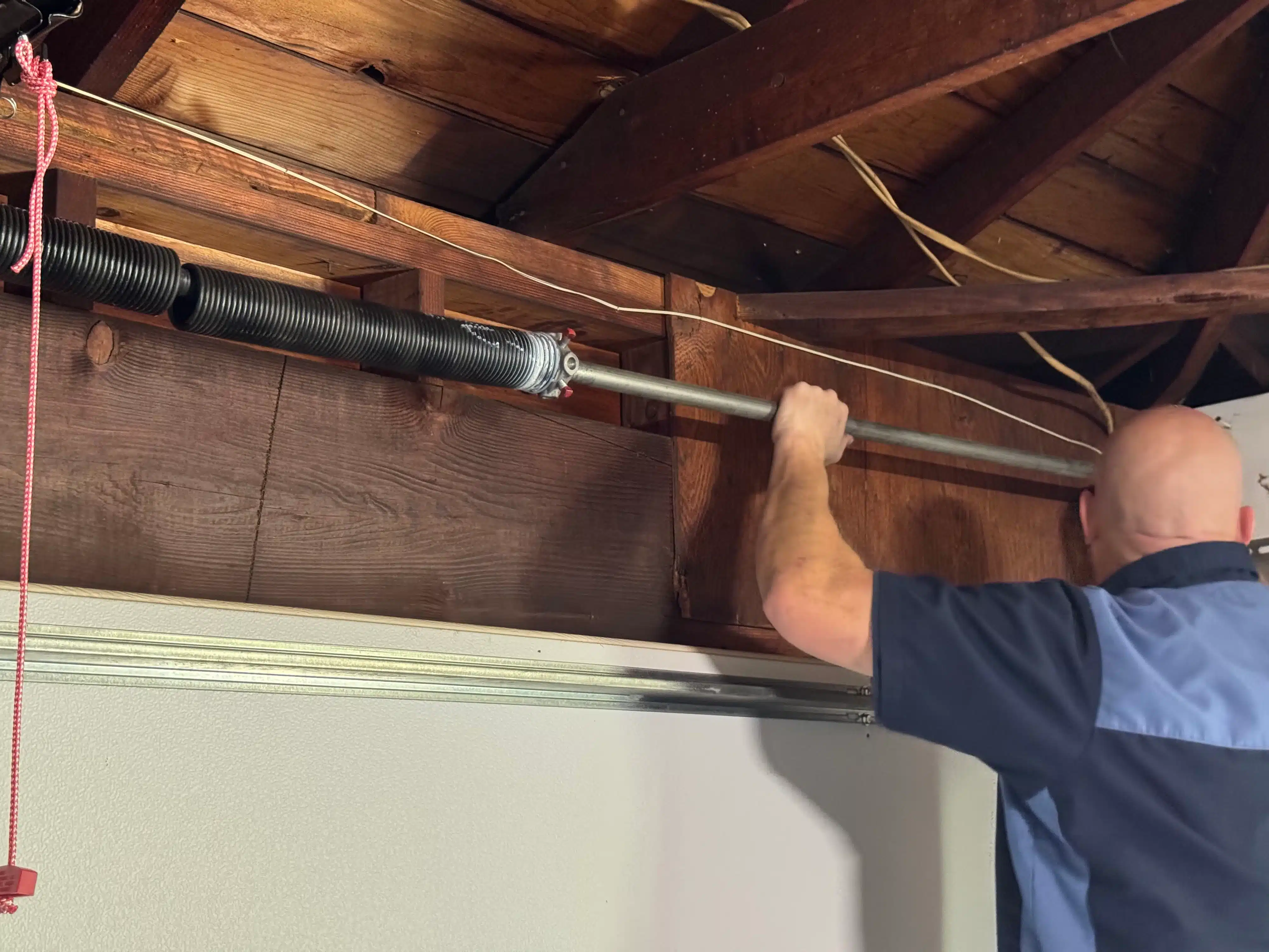 A service technician inspects and adjusts a heavy-duty metal torsion shaft assembly fixed against a wooden structural header, highlighting why hardware is best handled and garage door springs replaced by professionals.