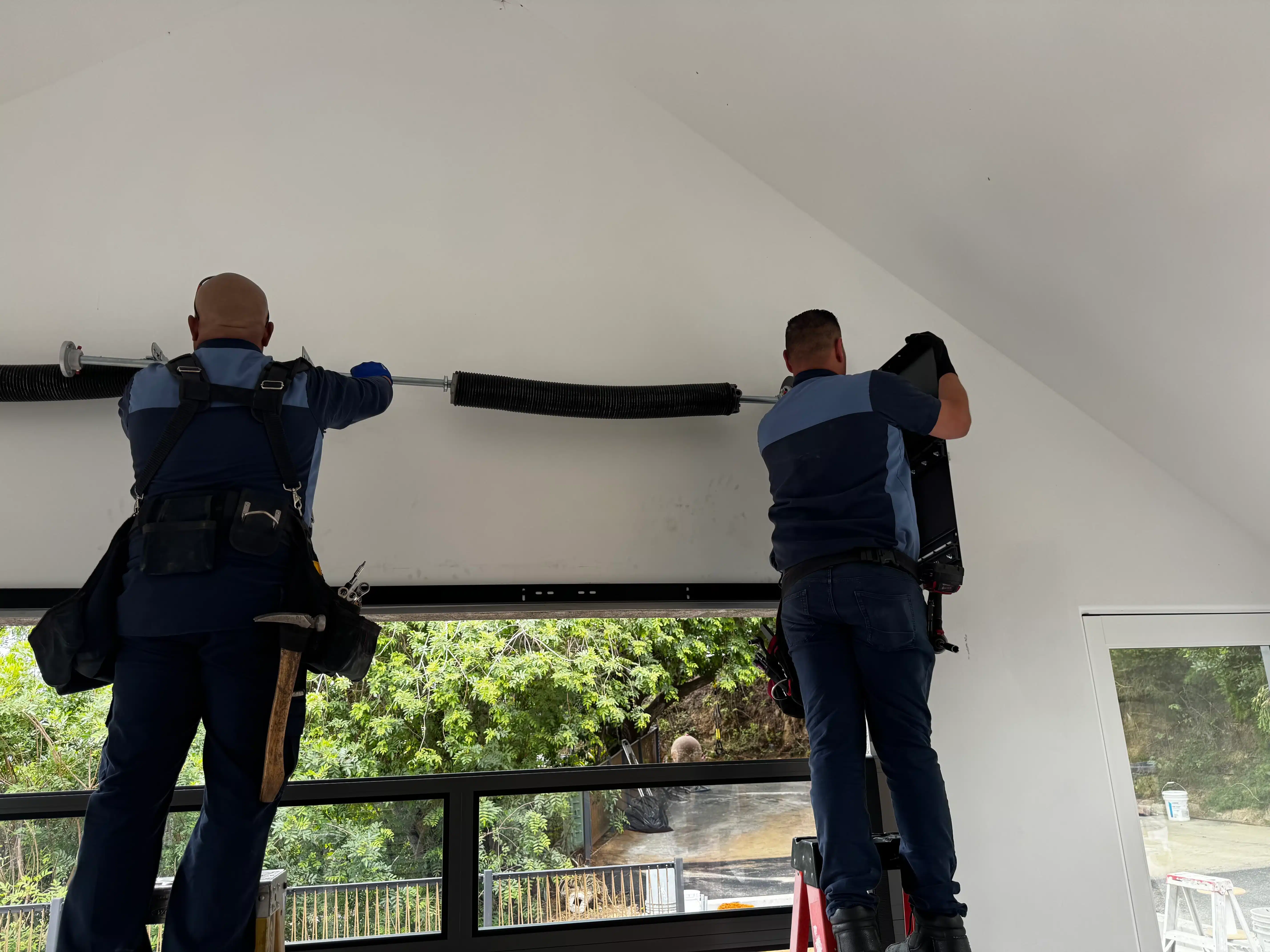 Two service technicians working together on scaffolding and a ladder to install and align an overhead dual torsion assembly, highlighting professional maintenance on how to keep your garage door springs in good shape.