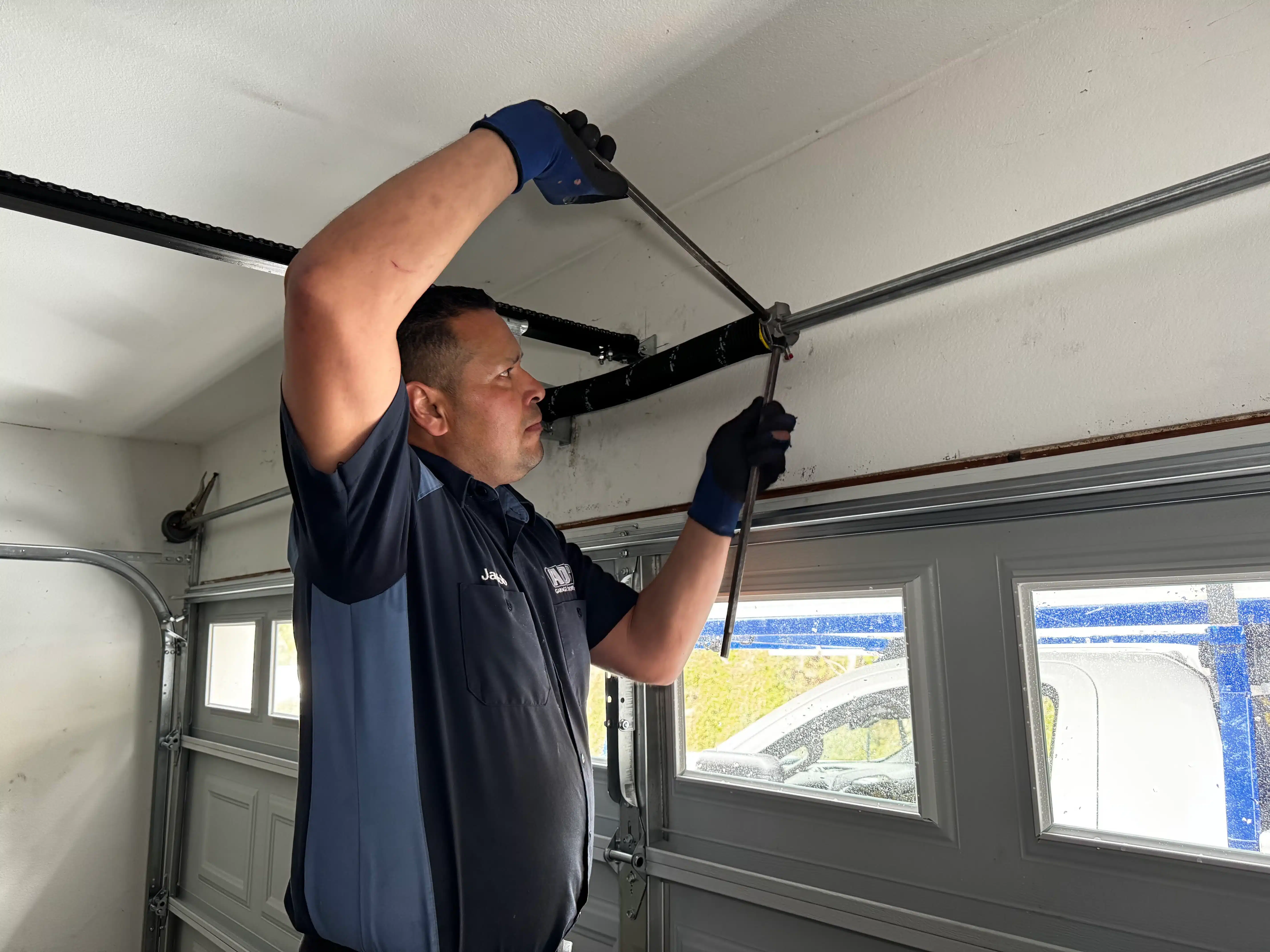 A professional service technician wearing safety gloves uses dual winding bars to safely tension an overhead torsion system, executing a professional garage door spring repair.